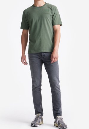 Olijfgroene T-shirt met korte mouwen in een zachte, gladde stof, gecombineerd met donkergrijze jeans en grijze sneakers, staande tegen een neutrale achtergrond.