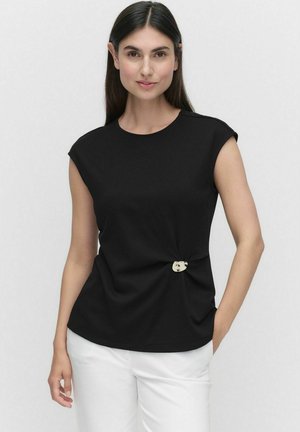 Black short-sleeve top with a round neckline, knot detail at the waist, and a decorative button accent; paired with white pants.