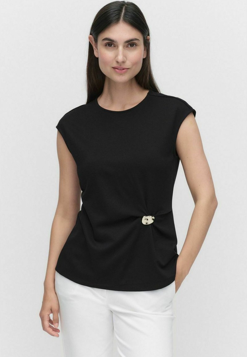 Zwarte korte mouwen top met een ronde halslijn, knoopdetail bij de taille en een decoratieve knopenaccent; gecombineerd met witte broek.