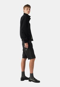 Giacca di jeans nera con chiusura a bottoni, abbinata a pantaloni shorts di jeans neri, calze grigie e mocassini neri. Il modello è in profilo su uno sfondo bianco.