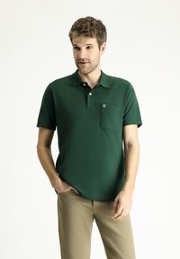 Polo verde con maniche corte, colletto con bottoni e tasca sul petto. Realizzato in tessuto liscio con una sottile trama.