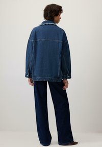 Persona stāv ar muguru pret kameru, ģērbusies pārmērīgi lielā tumši zila denima jakā un platajās biksēs uz vienkārša fona.