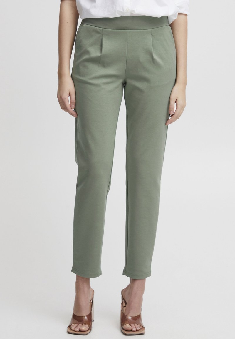 Femme portant un pantalon slim vert olive et des talons ouverts marron, debout devant un fond clair uni.