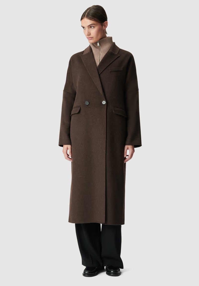 Bruine wollen jas met een lange, rechte silhouet, dubbele knopen en grote zakken; gecombineerd met een beige coltrui en zwarte broek.