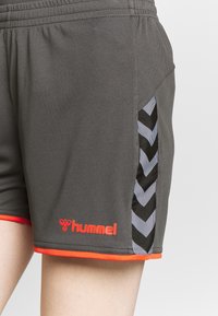 Pantalones cortos deportivos grises con paneles laterales en patrón de chevron en blanco y negro, ribete naranja y un logo rojo en la parte inferior izquierda. Hechos de tela ligera.