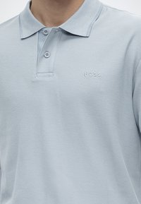 Polo bleu clair avec deux boutons et "BOSS" brodé sur la poitrine gauche, porté par une personne avec un cou visible et une épaule partielle.