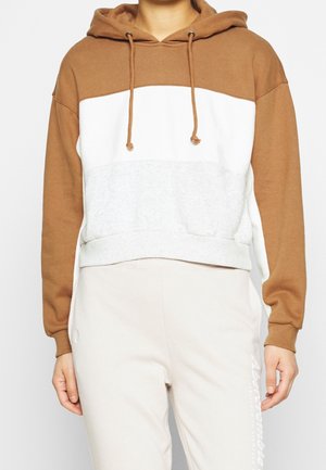 Person trägt einen Color-Block-Hoodie mit braunen Ärmeln und Kapuze, weißen und hellgrauen Bereichen am Körper, kombiniert mit hellen Hosen.