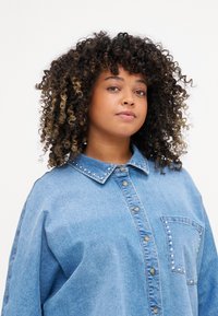 Camisa de ganga em azul claro, com colarinho clássico, fecho à frente com botões e um bolso com detalhes de tachas ao longo da costura. Cabelo encaracolado visível.