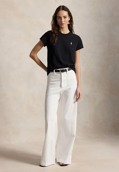 Γυναίκα που φοράει μαύρο πουκάμισο με κοντό μανίκι και λογότυπο, λευκό παντελόνι ψηλόμεσο με φαρδιές μπλούζες, μαύρη ζώνη και μαύρα παπούτσια ανοιχτά στη μύτη, στέκεται με το χέρι στη μέση.