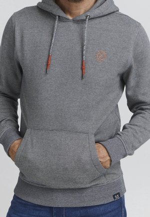 Ein Mann trägt einen grauen gestreiften Hoodie mit Fronttasche und Kordelzügen, Hände in den Taschen, kombiniert mit blauen Jeans.
