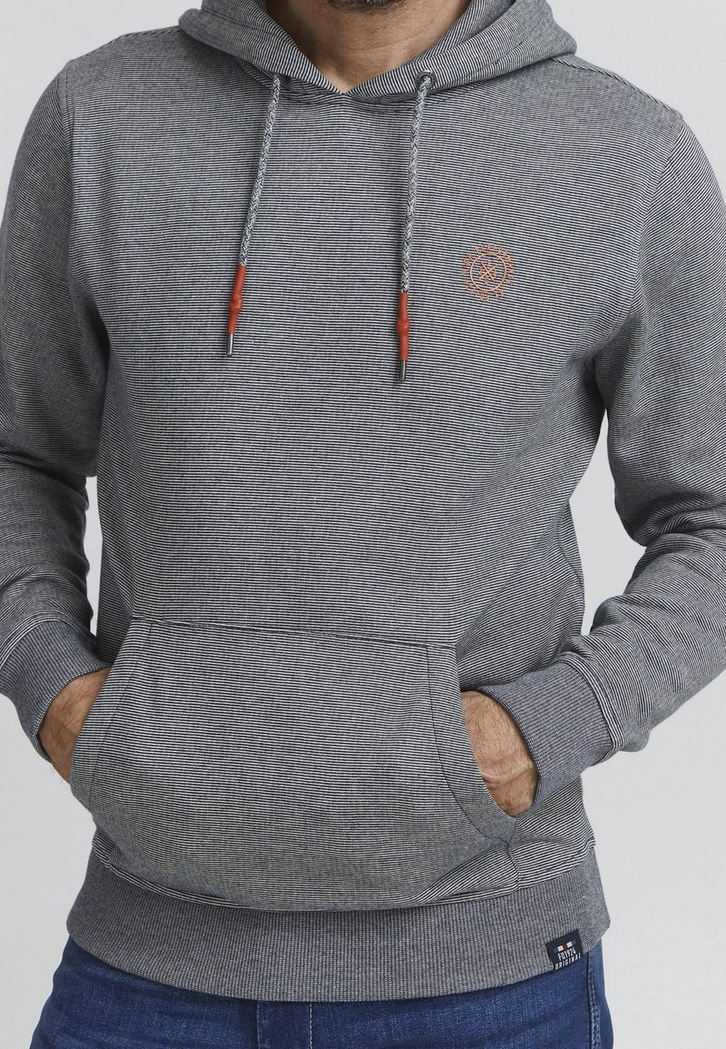 Homme portant un sweat à capuche gris rayé avec poche avant et cordons, mains dans les poches, associé à un jean bleu.