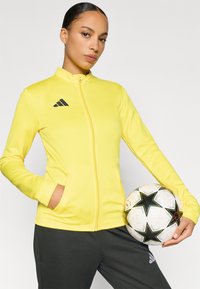 Κίτρινο αθλητικό μπουφάν με φερμουάρ και πλαϊνές τσέπες, με λογότυπο της Adidas. Το μοντέλο κρατά μια μπλούζα ποδοσφαίρου με μαύρα και άσπρα σχέδια.