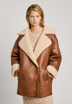 Cappotto doppiopetto in pelle marrone con colletto in shearling beige, vestibilità over e tasche laterali. Maglione di maglia crema indossato sotto.