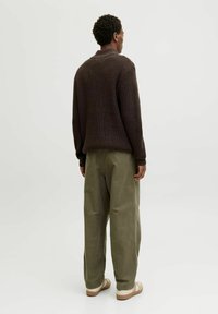 Maglione marrone lavorato a maglia con polsini a coste, abbinato a pantaloni larghi verde oliva. Il modello indossa scarpe beige slip-on. Sfondo minimalista.