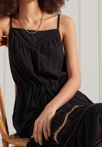 Svart, ärmlös klänning med elastisk midja och justerbar knytning, med texturerad tyg och subtila mönster samt en guldig accentkant.