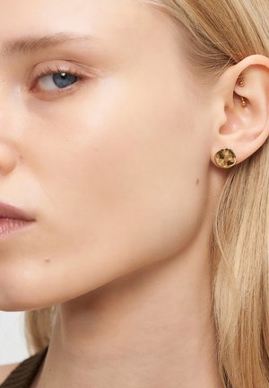 Close-up van het linkeroor van een vrouw met twee gouden oorbellen: een ronde stud op de oorlel en een kleine balpiercing in het kraakbeen van het oor.