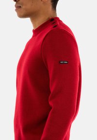Maglione rosso lavorato a maglia con scollo rotondo, dotato di tre bottoni sulla spalla e di un piccolo marchio "SAINT JAMES" sulla manica.