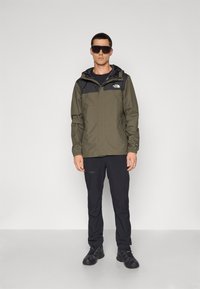 Waterdichte olijfgroene jas met zwarte accenten, ritsluiting, capuchon en verstelbare manchetten, gecombineerd met zwarte broek en zwarte schoenen.