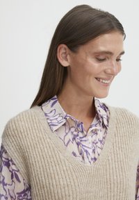 Gilet a coste beige in maglia sovrapposto a una blusa floreale lilla con colletto. I tessuti sembrano morbidi, con trame e motivi distintivi.