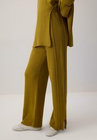 Ribidega, sinepikollane kudum, mille juurde kuuluvad laiad püksid, millel on külgsed lõhikuid ja nööpide detail ülal.