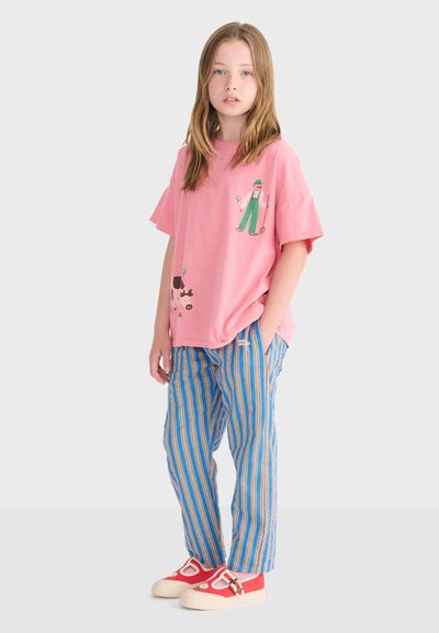 Młoda osoba stoi, ubrana w różowy, za duży T-shirt z nadrukiem rolnika, niebieskie spodnie w paski oraz czerwone buty z detalem stokrotki.