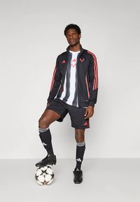 Schwarze Sportjacke mit roten Streifen, passende Shorts und ein weißes Shirt mit schwarzen Streifen; steht auf einem schwarz-weißen Fußball.