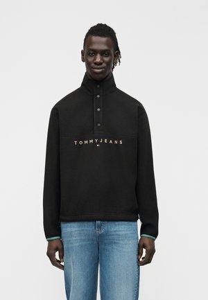 Moški s kitkami, oblečen v črno Tommy Jeans pulover s srajčnim ovratnikom na gumbe in svetlo modre kavbojke, stoji pred enostavnim ozadjem.