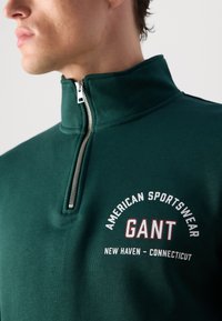 Zbliżenie na osobę noszącą ciemnozieloną bluzę z kapturem na zamek, na której piersi znajduje się napis "American Sportswear GANT New Haven - Connecticut".