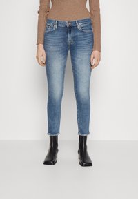 Jeans ajustados de mezclilla azul claro con un acabado desgastado, diseño de cinco bolsillos y cierre de botón, combinados con botas negras hasta el tobillo.