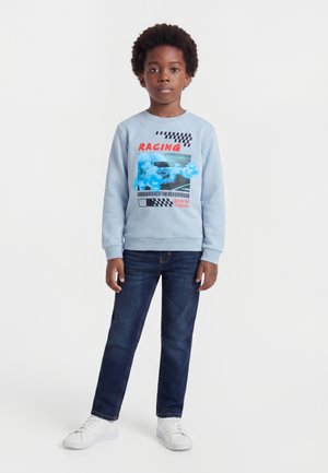 Barn iført en lyseblå sweatshirt med racerbils grafik og mørke jeans, stående med et neutralt udtryk og hvide sneakers.