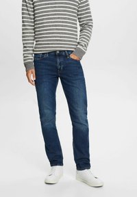 Jean en denim bleu foncé à coupe droite. Un homme porte un pull gris à rayures et des baskets blanches, les mains dans les poches.