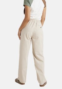Femme portant un pantalon large beige à rayures avec taille élastique, une chemise bicolore à manches courtes et des chaussures marron sans lacets, debout devant un fond blanc.
