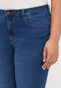 Blå denimjeans med klassisk snitt, gullknapplukking, fem lommer og synlige sømdetaljer. Myk tekstur.