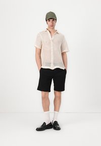 Crème kleurige mesh knoop shirt met korte mouwen, gecombineerd met zwarte shorts, witte sokken en zwarte schoenen, en afgewerkt met een groene pet.