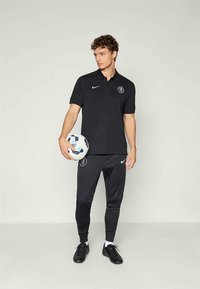 Črn Nike polo majica z logojem, v kombinaciji s črnimi hlačami in črnimi čevlji. Drži nogometno žogo in stoji pred nevtralno ozadje.