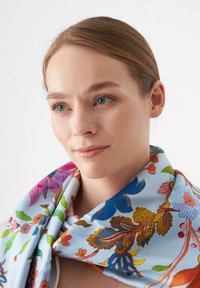 Écharpe en soie avec un fond bleu clair, présentant un motif floral vibrant dans des nuances de rose, vert, orange et bleu, avec une texture lisse.
