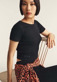 Top nero a maniche corte con design aderente, caratterizzato da un dettaglio con logo. La modella indossa una sciarpa con stampa leopardata in vita.