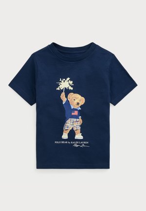 Námornícke bavlnené tričko s grafikou medveďa v modrom saku, s americkou vlajkou, károvanými šortkami a bielymi teniskami. Značka Ralph Lauren je zahrnutá.