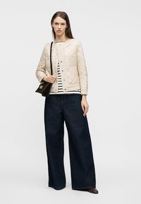 Γυναίκα που φοράει μπεζ μπουφάν με καπιτονέ λεπτομέρειες, ριγέ πουκάμισο, ευρύχωρο σκούρο τζιν παντελόνι, καφέ παπούτσια και κρατά μια σκούρα καφέ τσάντα ώμου.