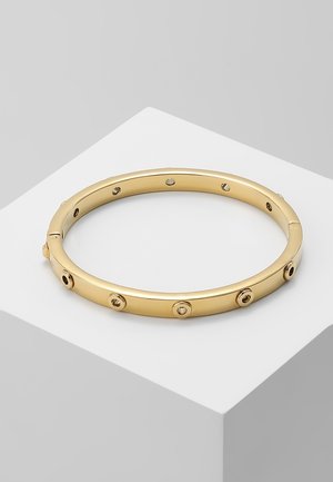 Gouden armband met regelmatig geplaatste ronde schroefmotieven, rustend op een wit geometrisch platform tegen een lichtgrijze achtergrond.