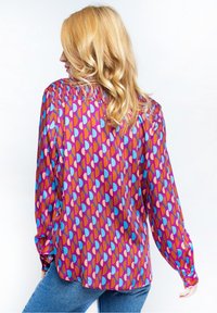 Blusa a maniche lunghe con base viola, caratterizzata da un motivo geometrico di forme rosa, blu e arancio. Dettagli arricciati sulle spalle.