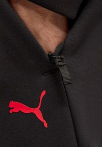 Schwarzes zip-tops mit strukturiertem Stoff, mit einem auffälligen roten Puma-Logo. Die Darstellung zeigt eine Nahaufnahme des Reißverschlusses und des Ausschnitts.