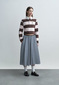 Nő áll egy egyszerű háttér előtt, barna és rózsaszín csíkos, rövid fazonú felsőt, kék-fehér csíkos hosszú szoknyát, fehér zoknit és sötét mokaszinokat visel.