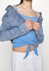 Džínová bunda s opotřebovanými detaily, světle modrý top uvázaný v pase a bílé kalhoty s vysokým pasem. Perlový náhrdelník dodává vzhledu detail.