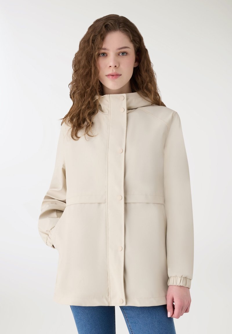 Giovane donna con capelli ricci castani, indossa un cappotto beige con bottoni, con le mani nelle tasche e jeans blu, in piedi contro uno sfondo bianco.