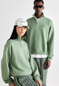 Green zip-up sweatshirts and patterned green pants, paired with a white cap featuring black text, against a neutral background.