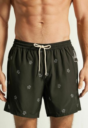 Männlicher Oberkörper mit schwarzen Badeshorts mit weißen Schildkrötenmustern und weißem Kordelzug, zeigt einen Flaschenöffner, der am Bund befestigt ist.