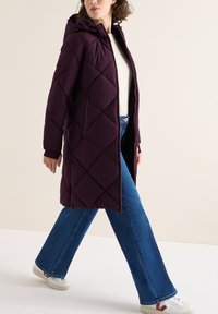 Lungo cappotto a palloncino viola con motivo a rombi, cappuccio e tasche laterali, abbinato a jeans svasati blu e sneakers bianche.