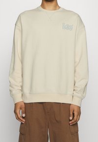 Beige mikina s kulatým výstřihem a dlouhými rukávy. Na hrudi je vyšitý modrý logo 'Lee'. Měkká, hladká textura.