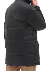 Person justerer en sort dunjakke bagfra, iført beige bukser, mod en simpel hvid baggrund.